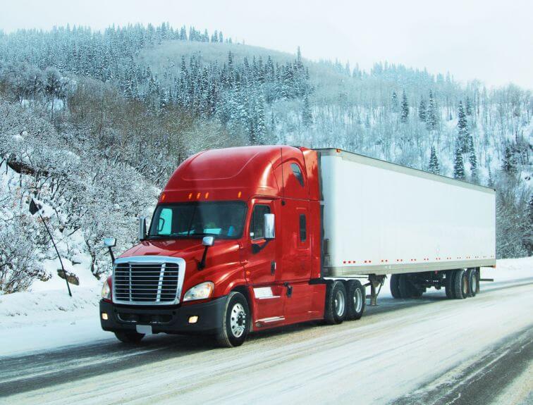 Image of red and white semi truck driving on a snow-covered road with a hill of trees in the background
