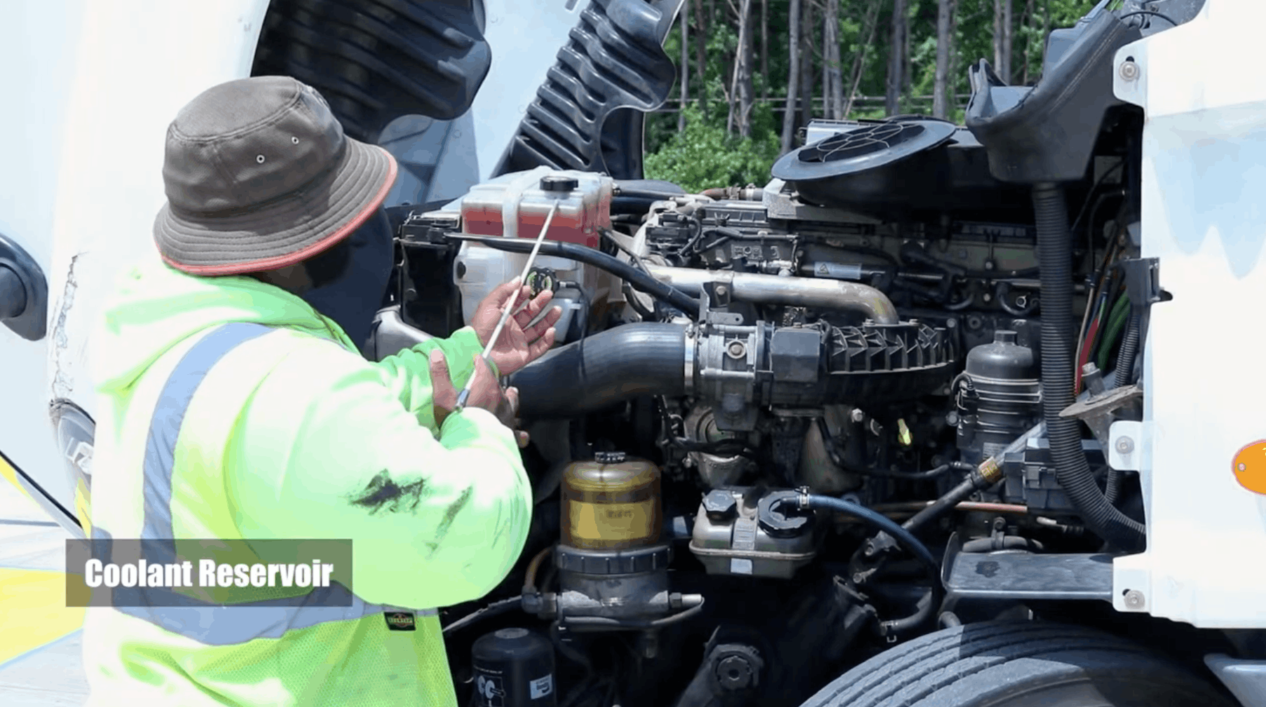 CDL instructor pointing at coolant reservoir in the hood of semi truck with a stick