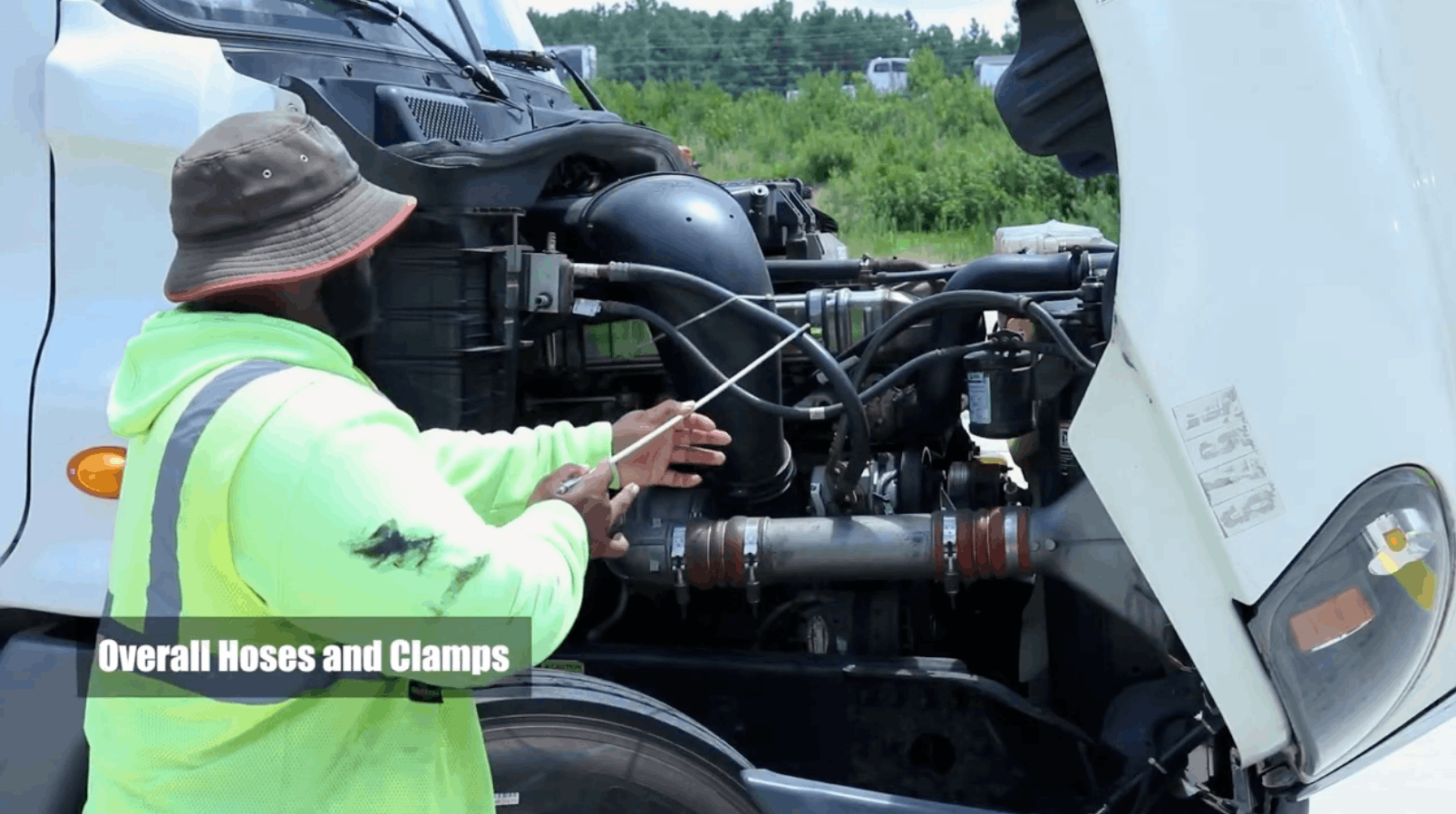 CDL instructor pointing to hoses and clamps with stick, inside front of semi truck