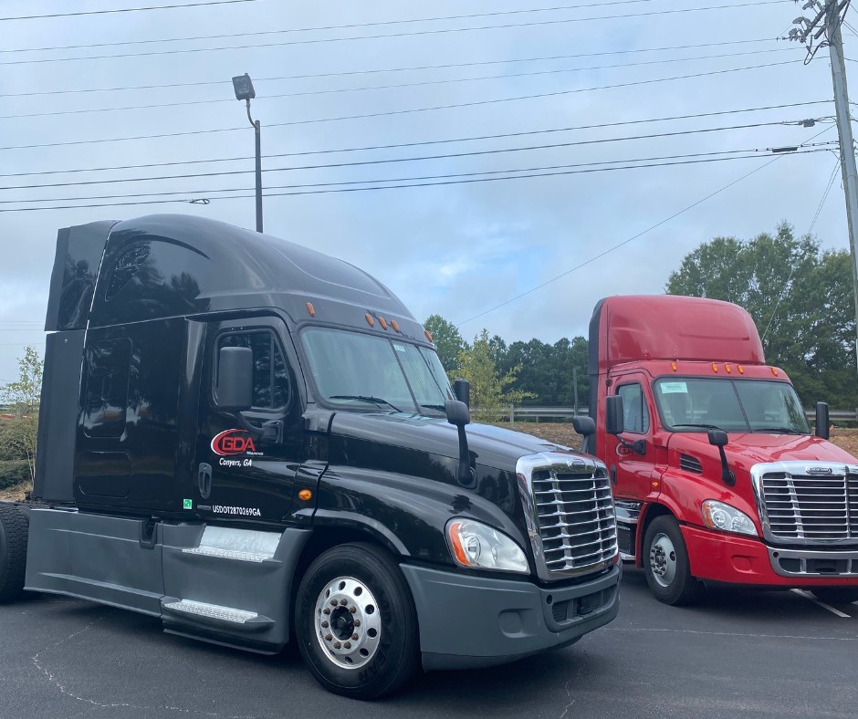 A black GDA training semi truck from Conyers, Georgia, parked beside a red day cab semi truck in a lot